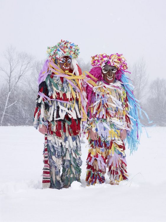 Zwei Personen im Schnee, die in bunte, fransige Kostüme gekleidet sind und grinsende Holzmasken tragen.