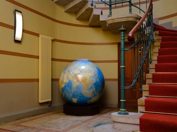 Großer Globus in einem alten Treppenhaus. Links von der Weltkugel ist eine Wandheizung zu sehen. Rechts führt eine Treppe mit rotem Teppich nach oben.