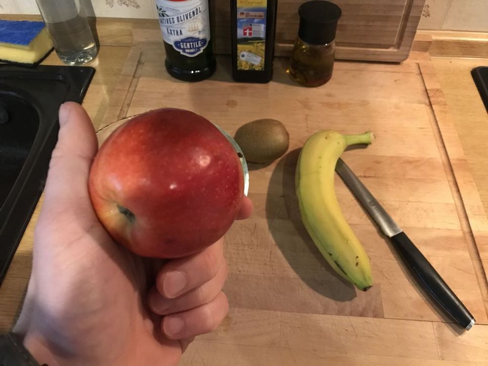 Eine Hand hält einen roten Apfel, eine Banane, eine Kiwi und ein Messer liegen auf einem Holzbrett.